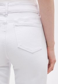 Κοντινό πλάνο ενός ατόμου που φορά ψηλόμεσα λευκά τζιν, με ορατή πίσω τσέπη και μέρος του δεξιού βραχίονα, πάνω σε απλό φόντο.