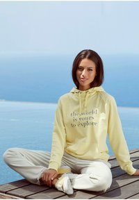 Geel hoodie met de grafische tekst "de wereld is van jou om te verkennen." Gecombineerd met lichtbeige broek en bijpassende schoenen, zittend op een houten dek.