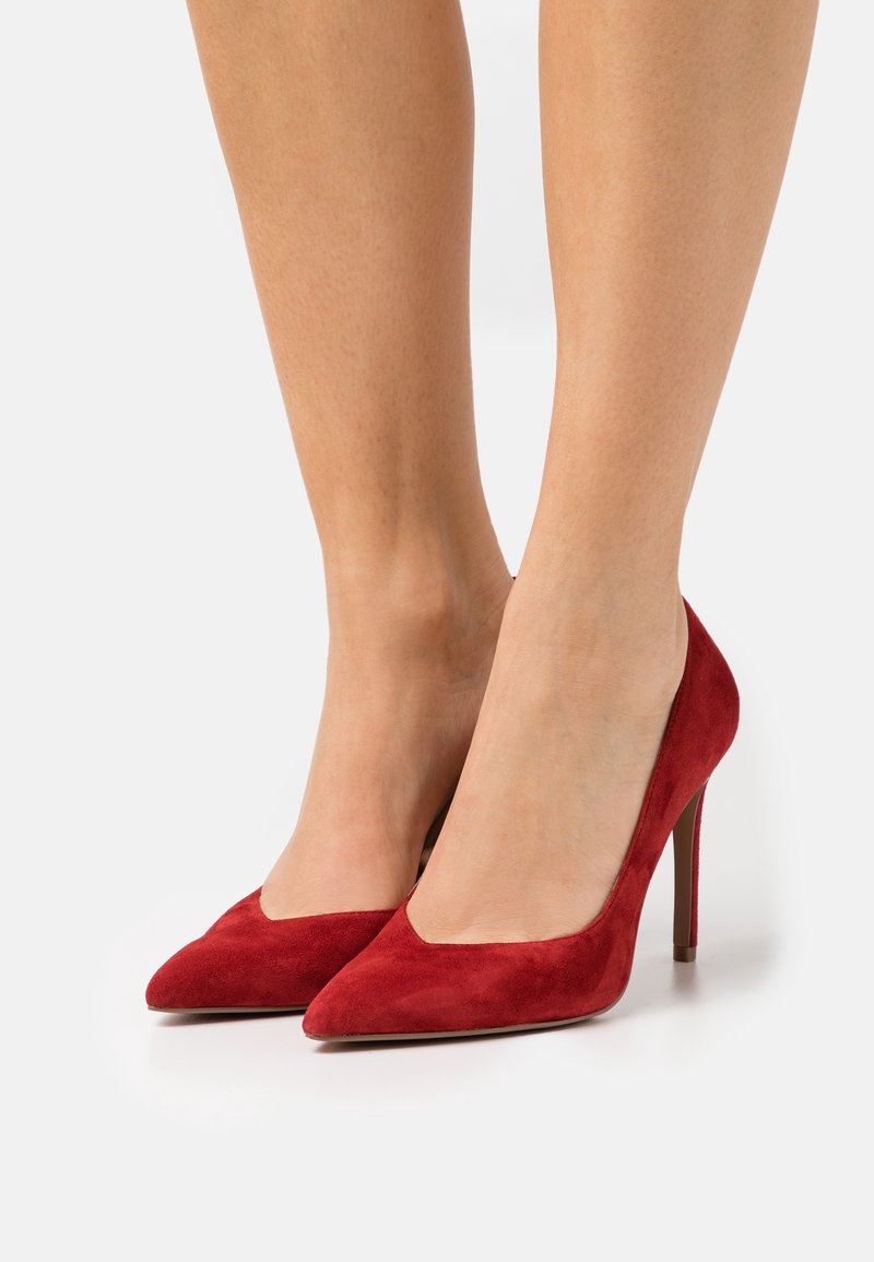 Femme portant des escarpins en suède rouge à bout pointu avec talons aiguilles fins, debout sur une surface blanche.