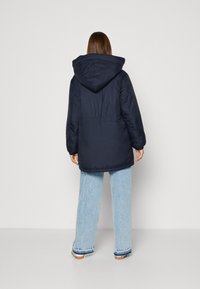 Giacca imbottita blu navy con cappuccio, polsini elasticizzati e vestibilità comoda. Indossata sopra jeans a gamba larga blu chiaro con rifiniture sull'orlo.