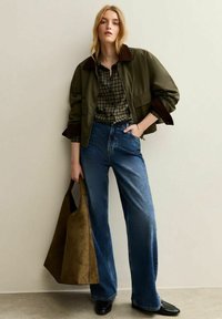 Giacca bomber verde oliva con colletto marrone, camicia a quadri, jeans blu a vita alta e borsa a tracolla in suede beige. Scarpe nere completano il look.