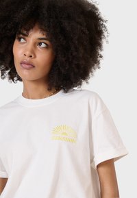 Weißes Baumwoll-T-Shirt mit gesticktem gelben Sonnenmotiv und dem Wort "SUNSHINE." Klassische Passform mit aufgerollten Ärmeln und weicher Textur.