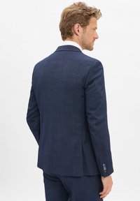 Mężczyzna widziany z tyłu, ubrany w ciemnoniebieską, wzorzystą marynarkę w komplecie z pasującymi spodniami oraz białą kołnierzykiem koszuli widocznym.