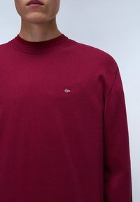 Suéter burdeos de manga larga con un cuello acanalado y un pequeño detalle de bandera bordado en el lado izquierdo del pecho. Textura de punto suave y corte relajado.