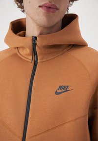 Sweat à capuche Nike orange avec une fermeture éclair noire et un logo, présentant un design ajusté et un tissu doux au toucher. Détails de couture visibles.
