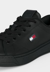 Baskets en cuir noir avec des lacets noirs, des œillets métalliques et un petit patch logo Tommy Jeans sur le côté.