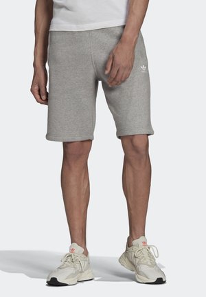 Mand iført lysegrå Adidas-shorts, hvide sportsko og en hvid skjorte, der står på en ensfarvet lys baggrund.