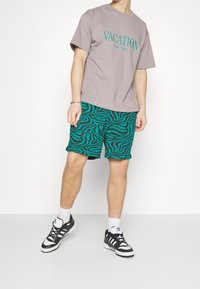 T-shirt gris avec le texte "VACANCES" en teal ; associé à un short vert teal avec un motif de zèbre noir ondulé. Baskets noires, chaussettes blanches.