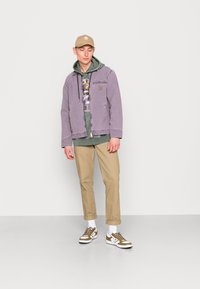 Giacca di jeans viola sopra un felpa verde, t-shirty grafica, chinos beige e sneakers marroni con dettagli bianchi. Outfit semplice e casual.