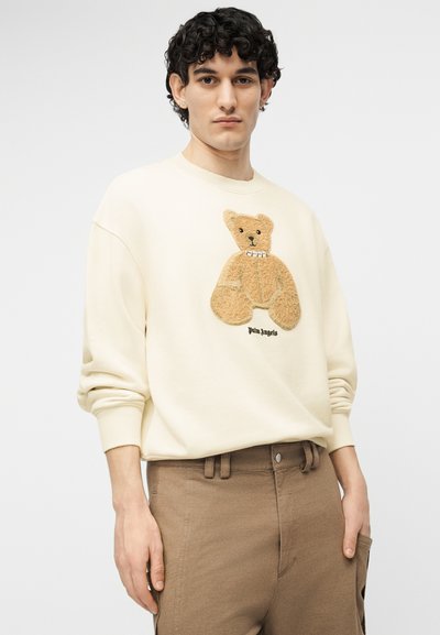 Νεαρός με σγουρά μαλλιά, που φοράει εκρού φούτερ με ανάγλυφη εικόνα αρκουδάκι teddy και καφέ παντελόνι, σε μονόχρωμο φόντο.