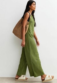Jumpsuit verde a righe con una vestibilità rilassata, abbinato a una borsa a tracolla in tessuto color tan e sandali bianchi. Presenta gambe larghe e design senza maniche.