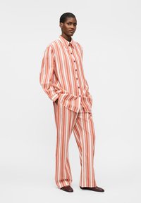 Femme debout les mains dans les poches, portant une chemise à rayures verticales orange et blanche ample et boutonnée, ainsi qu’un pantalon large assorti avec des chaussures foncées.