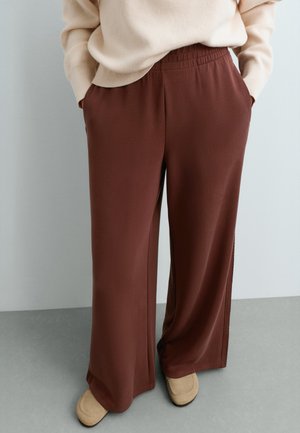 Person som bär lösa bruna byxor med elastisk midja, beige topp och beige skor, stående med händerna i fickorna.