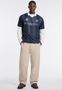 Granatowa koszulka polo z białym kołnierzykiem, z poziomymi paskami i napisem "GUESS JEANS". W komplecie z beżowymi spodniami i brązowymi butami.