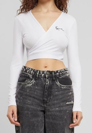 Nainen pukeutunut valkoiseen pitkähihainen midja-pituisella kietaisutopilla, jossa on musta logodetalji, ja korkeavyötäröisiin haalistuneisiin mustiin farkkuihin, joissa on kulunut kohta.