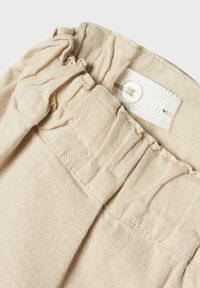Beige linnetyg med samlad textur vid midjan. Har en knappstängning och en tygetikett med ett sytt mönster.