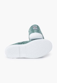 Zapatillas de lona verde con suelas de goma blanca y patrones texturizados. Presentan una etiqueta cosida en el talón y un diseño de punta redondeada.