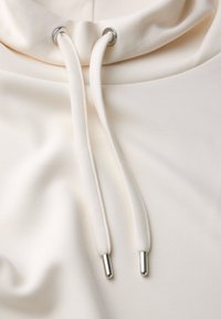 Light cream hoodie featuring a high neckline, silver eyelets, and drawstrings with metal tips, made from a smooth, stretchy fabric.
