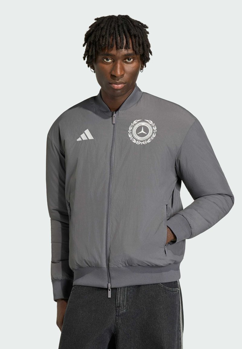 Homme portant une veste zippée grise avec les logos Adidas et Mercedes-Benz, debout avec une main dans la poche devant un fond uni.