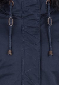 Chaqueta azul marino en la parte delantera con tapa de cremallera vertical, costuras en zigzag y dos cordones trenzados con puntas de metal y ojetes metálicos ovalados.