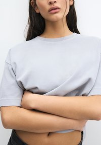 T-shirt gris clair à col rond, à manches courtes, au tissu doux, avec une coupe décontractée, porté avec les bras croisés, mettant en valeur une légère taille au-dessus de la taille.