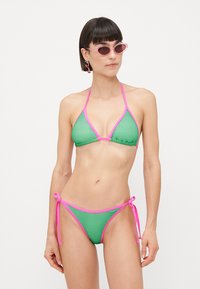 Groen mesh bikini met roze afwerking, voorzien van een driehoek-top en een zijknoop-bottem. Zonnebril met een roze tint, wat een contrasterend detail toevoegt.