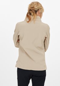 Beige mjukskaljacka med hög krage, som har sömmar och en något avsmalnande passform. Tyget verkar slätt och flexibelt.