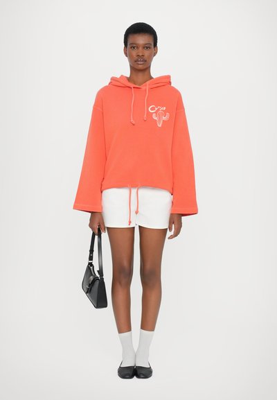 Korallfarbener Oversized-Hoodie mit einem weißen Kaktusdesign, kombiniert mit weißen Shorts. Die Accessoires umfassen eine schwarze Handtasche und graue Socken mit schwarzen Ballerinas.