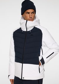 Herrjacka med en marinblå och vit färgschema; har ett vadderat bröst, hög krage och fickor med dragkedja med kontrasterande metalldetaljer.