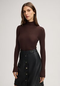 Brauner, gerippter Rollkragenpullover mit langen Ärmeln, kombiniert mit einem schwarzen Lederrock, der goldene Knopfakzente und eine taillierte Silhouette aufweist.