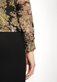 Blusa trasparente a fiori nei toni di verde, marrone e nero, con maniche a sbuffo con polsini elasticizzati, abbinata a un pantalone a tessuto nero aderente.
