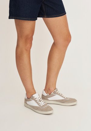Persoon die beige en witte sneakers met veters draagt en donkeren spijkershorts, staand op een lichte achtergrond.
