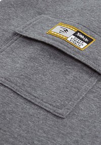 Grijze sweatshirtstof met een grote platte zak en een rechthoekig label met gele en zwarte tekst dat "Speed Strength Stealth" leest.