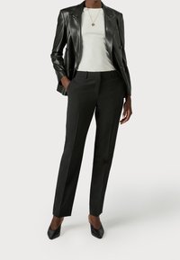 Woman wearing black leather blazer, white top, black tailored pants, black pointed-toe heels, and a gold pendant necklace against plain background.