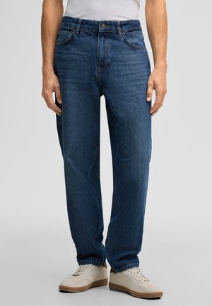 Mann trägt dunkelblaue Jeans mit geradem Bein und weiße Turnschuhe, steht vor einem schlichten hellgrauen Hintergrund.