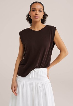 Kvinde med krøllet hår iført gyldne hoop-øreringe, sort ærmeløs top og hvid nederdel, poserer med hånden på hoften mod en ensfarvet baggrund.