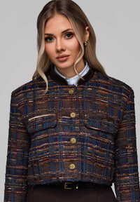Obcisła marynarka z tweedu w odcieniach niebieskiego, pomarańczowego i brązowego. Posiada kołnierz, złote guziki oraz dwie przednie kieszenie z kontrastowym wykończeniem.