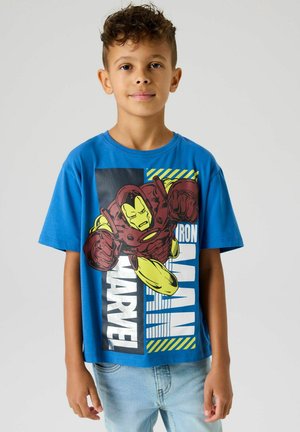 Jongen met kort, krullend haar, gekleed in een blauw T-shirt met een grote Iron Man Marvel-afbeelding, staande tegen een effen lichte achtergrond.