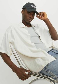 Hvid kortærmet skjorte med knapper, grå ribbet tanktop, blå denim jeans og sort kasket med teksten "NINETY", der sidder på en krom stol.