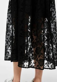 Jupe en dentelle noire avec un motif floral et des sections transparentes. L'ourlet est solide, avec un bord légèrement festonné. Assortie à des chaussures gris clair.