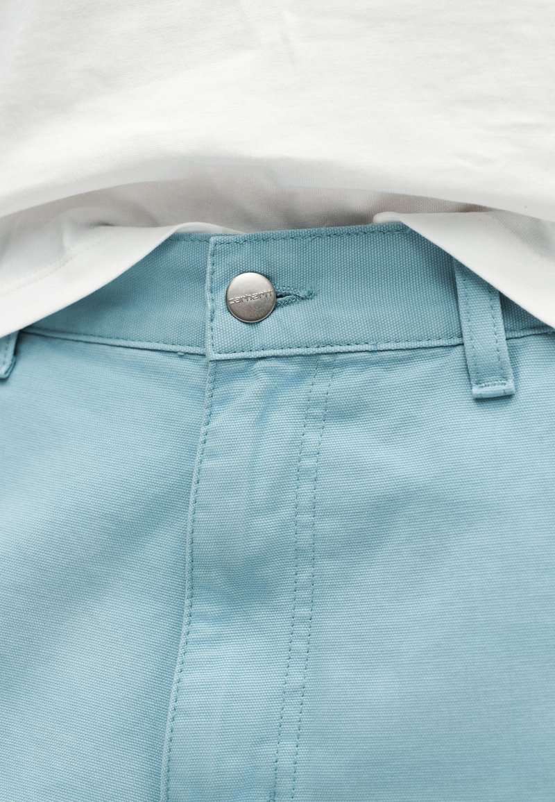 Pantalon bleu clair avec bouton visible et passants de ceinture, associé à un haut blanc rentré dans le pantalon au-dessus de la taille.