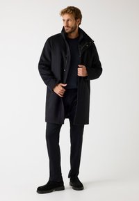 Cappotto in misto lana nero con collo alto e chiusure a pressione, indossato sopra un maglione scuro e pantaloni neri su misura, abbinato a mocassini neri.
