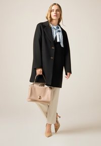 Abrigo de lana negro con botones dorados, corbata de estampado azul claro, pantalones crema, bolso beige y tacones puntiagudos a juego.