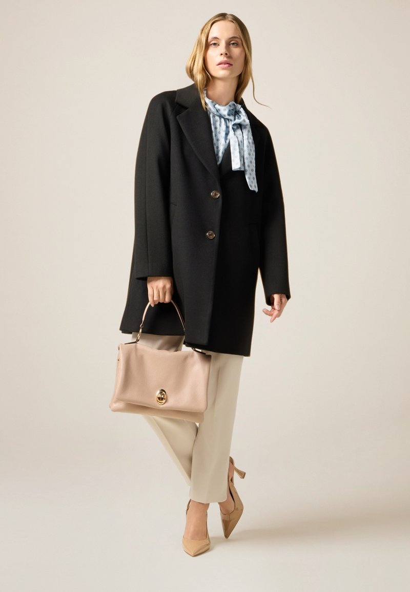 Abrigo de lana negro con botones dorados, corbata de estampado azul claro, pantalones crema, bolso beige y tacones puntiagudos a juego.