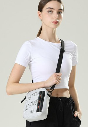 Jeune femme portant un t-shirt blanc court et un pantalon noir, tenant un sac bandoulière blanc imprimé avec une sangle noire sur l'épaule.