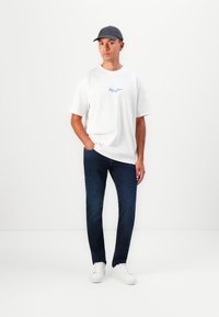 Weißes Baumwoll-T-Shirt mit blauer Schrift, dunkelblaue Jeans und weiße Sneakers. Das Model trägt eine dunkelgraue Baseballmütze.