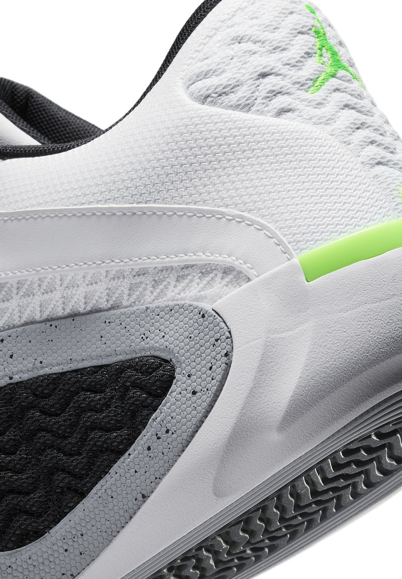 Zapatilla blanca con acentos grises y un panel texturizado negro. Presenta un logo verde, parte superior de malla y suela con patrón para mayor tracción.