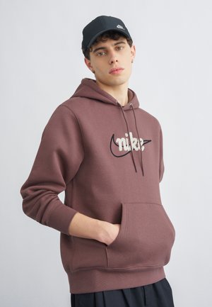 Mladý muž v hnedej mikine Nike s predným vreckom a čiernou šiltovkou Nike stojaci pred jednoduchým svetlým pozadím.