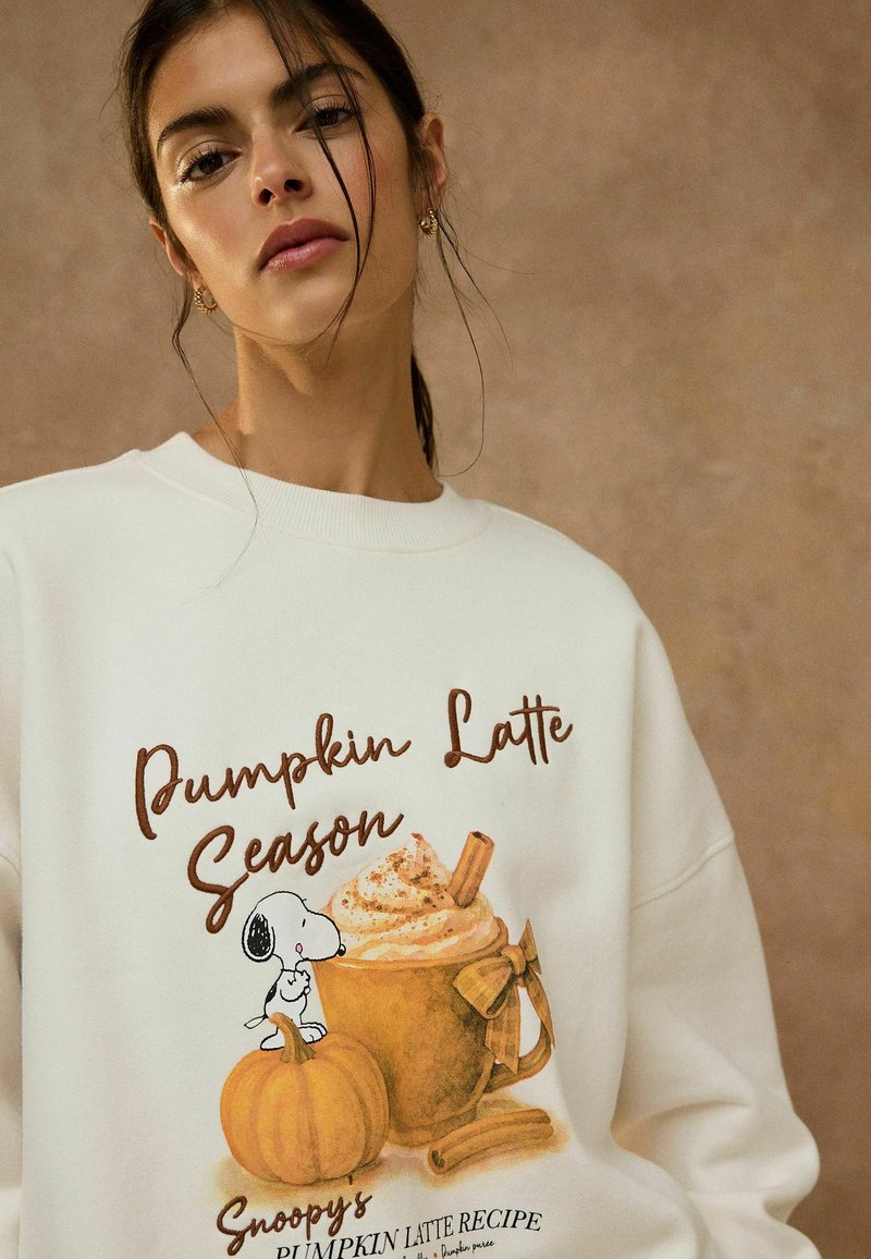 Young woman wearing a white sweatshirt with a "Pumpkin Latte Season" design featuring Snoopy, a pumpkin, and a whipped cream-topped latte.