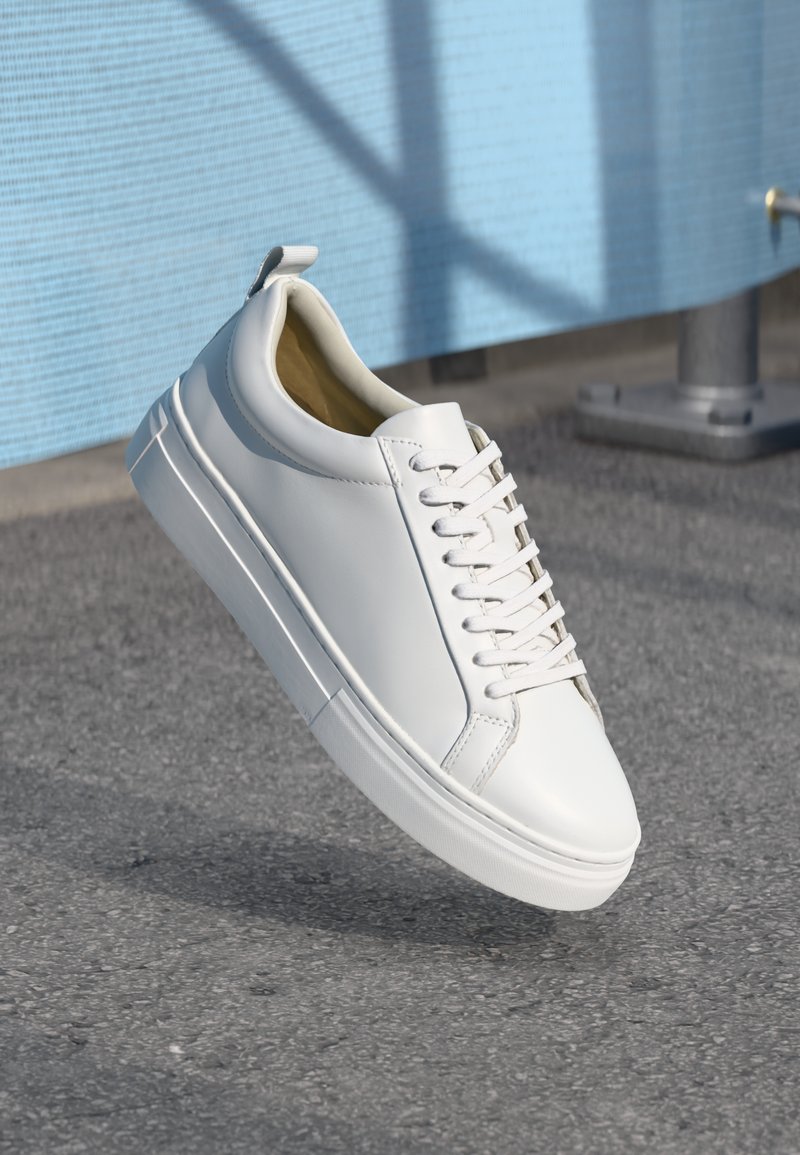 Sneaker bianco in pelle con lacci piatti, punta arrotondata, suola in gomma e una piccola linguetta sul tallone. Texture liscia con dettagli di design minimi.